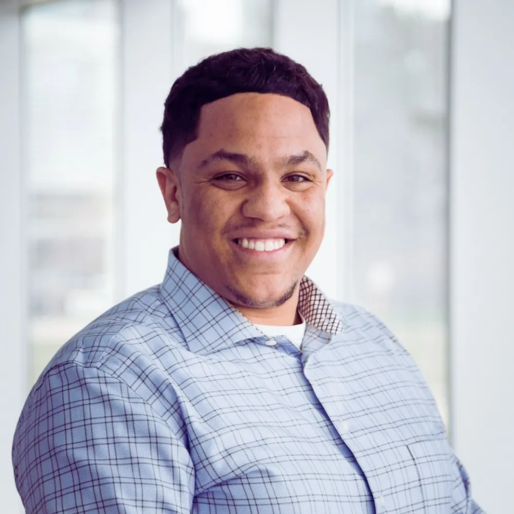 Aziz Abdullah, a dark skinned man smiling in a plaid button up shirt.