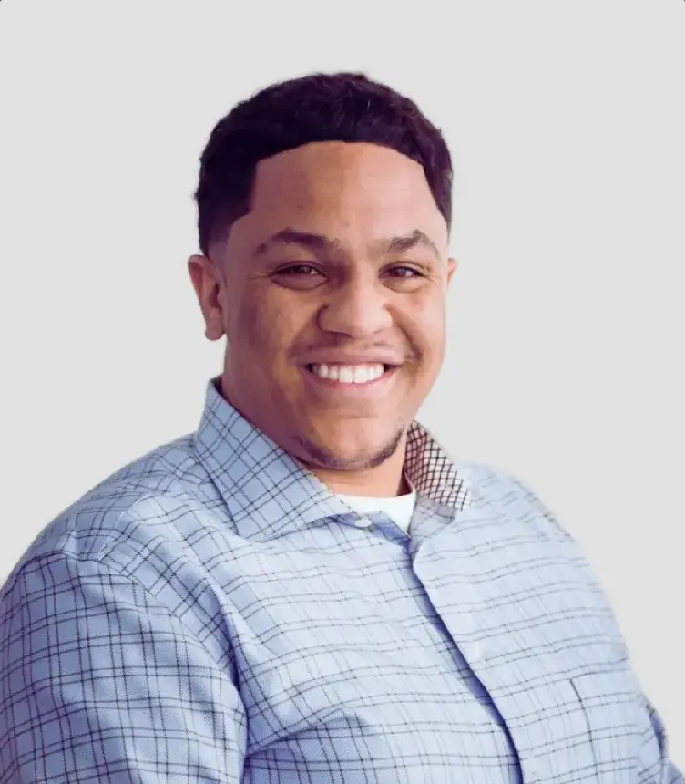 Aziz Abdullah, a dark skinned man smiling in a plaid button up shirt.